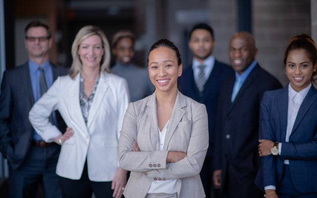 A group of colleagues stands as a collective in business attire. They are all smiling and very professional.